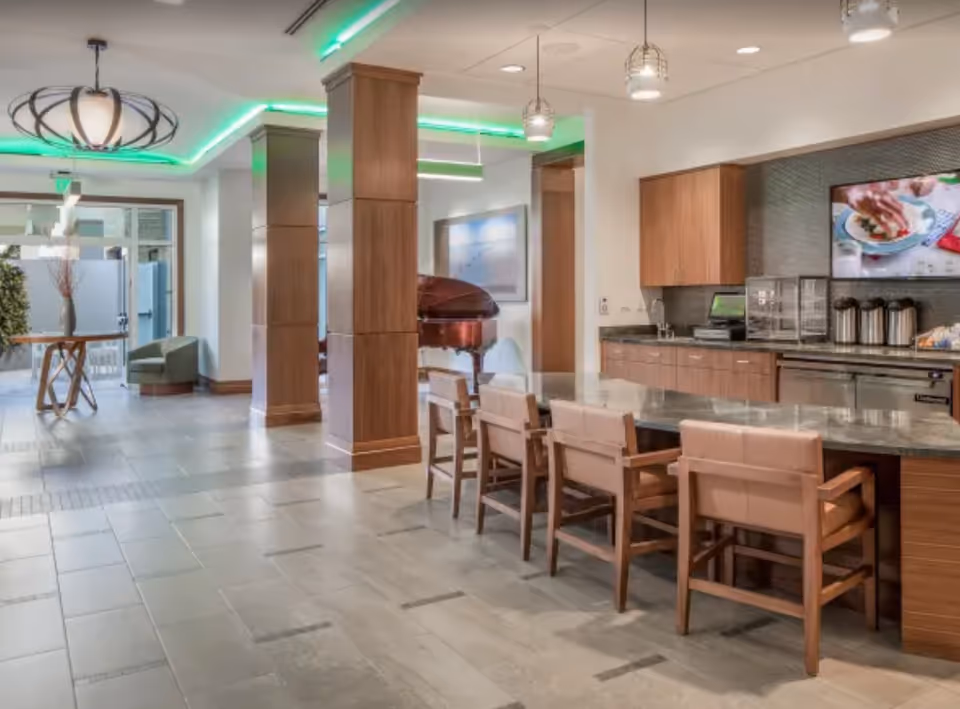 A modern common area in a senior living facility featuring a long counter with six cushioned wooden chairs, wooden cabinets, a countertop with coffee dispensers, and a wall-mounted TV displaying a cooking show. The room has tiled flooring, wooden pillars, and green accent lighting on the ceiling. In the background, there is a grand piano and a small seating area with a round table and two chairs near glass doors.