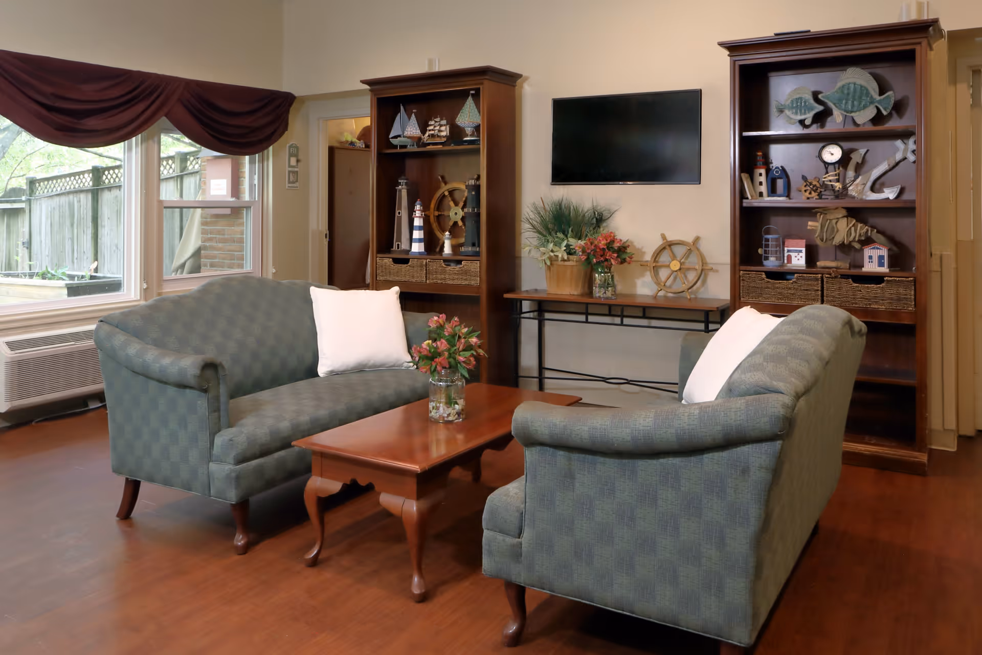 A cozy living room area with two green upholstered sofas facing each other across a wooden coffee table with a flower vase. Behind the sofas are two wooden bookshelves decorated with nautical-themed items such as model ships, a ship wheel, and fish sculptures. A flat-screen TV is mounted on the wall above a console table with plants and flowers. Large windows with maroon valances let in natural light, and the floor is wooden.
