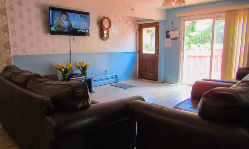 A cozy living room with two dark brown leather sofas facing a wall-mounted flat screen TV. The wall has floral wallpaper on the upper half and blue paint on the lower half. A wooden clock is mounted on the wall next to a wooden door with a window. There is a sliding glass door leading outside, allowing natural light to brighten the room. A small table with yellow tulips is placed near the TV.