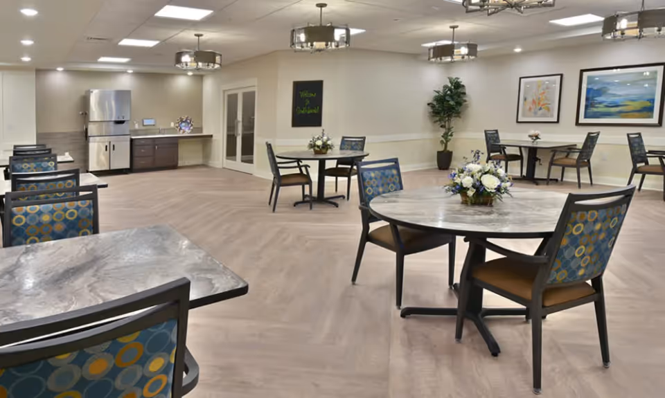 Spacious communal dining room with round and square tables, patterned chairs, floral centerpieces, and a serving area in the back.
