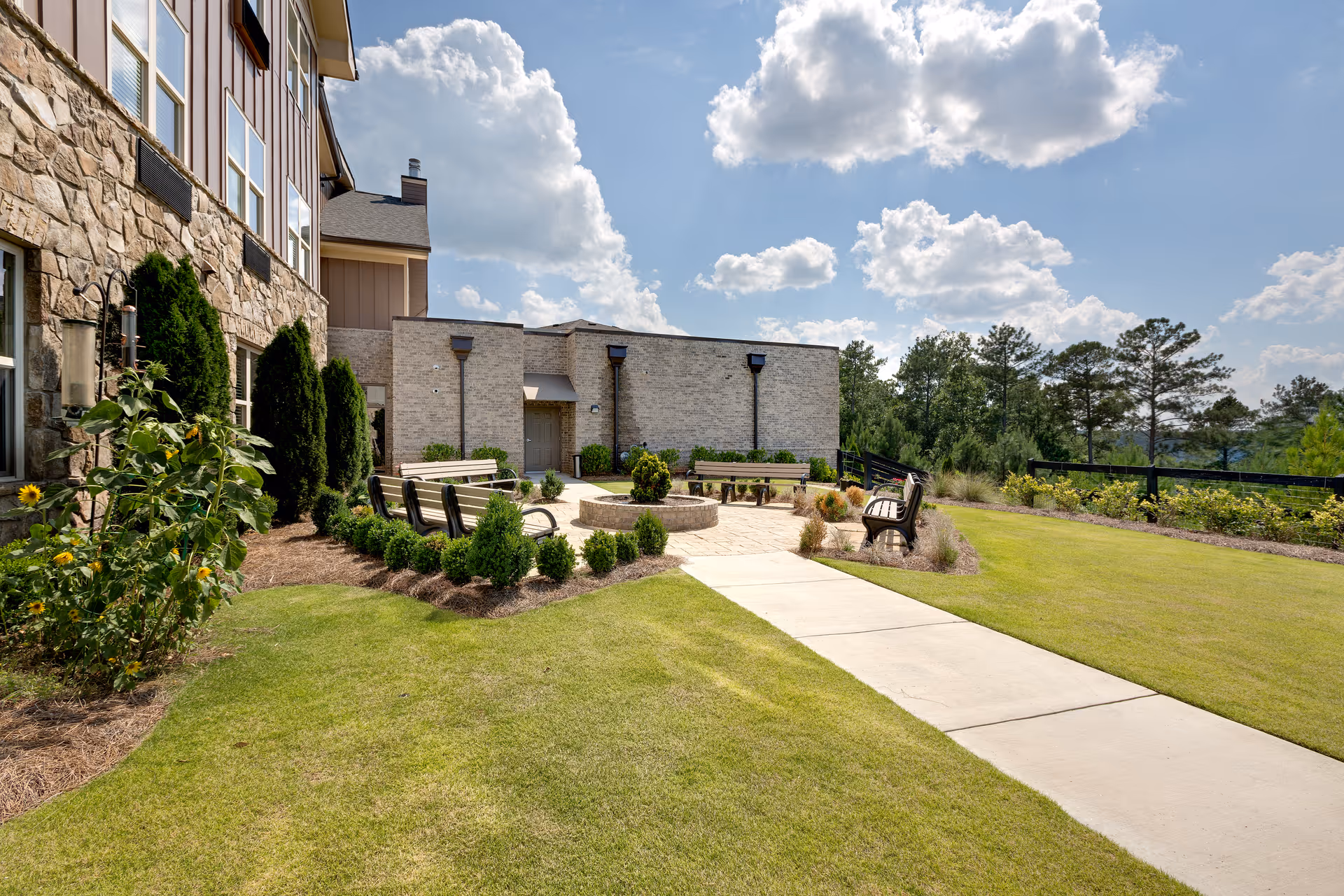 Outdoor seating area at Oaks at Acworth senior living facility with benches arranged around a circular fire pit, surrounded by well-maintained grass, shrubs, and trees under a partly cloudy sky.