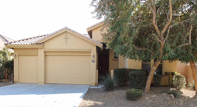 Exterior front view of a single-story beige stucco house with a tiled roof, two garage doors, a driveway, and a small front yard with trees and shrubs.