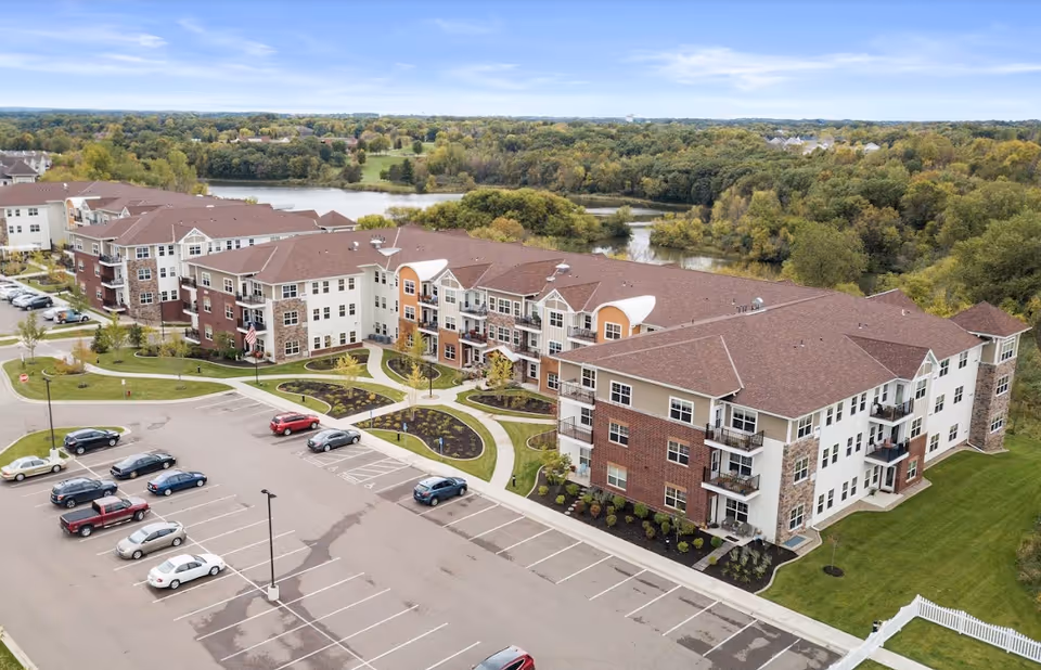Aerial view of a large senior living facility named Chaska Heights, featuring a multi-story building with balconies, surrounded by landscaped gardens and a parking lot with several cars. In the background, there is a wooded area and a body of water under a partly cloudy sky.