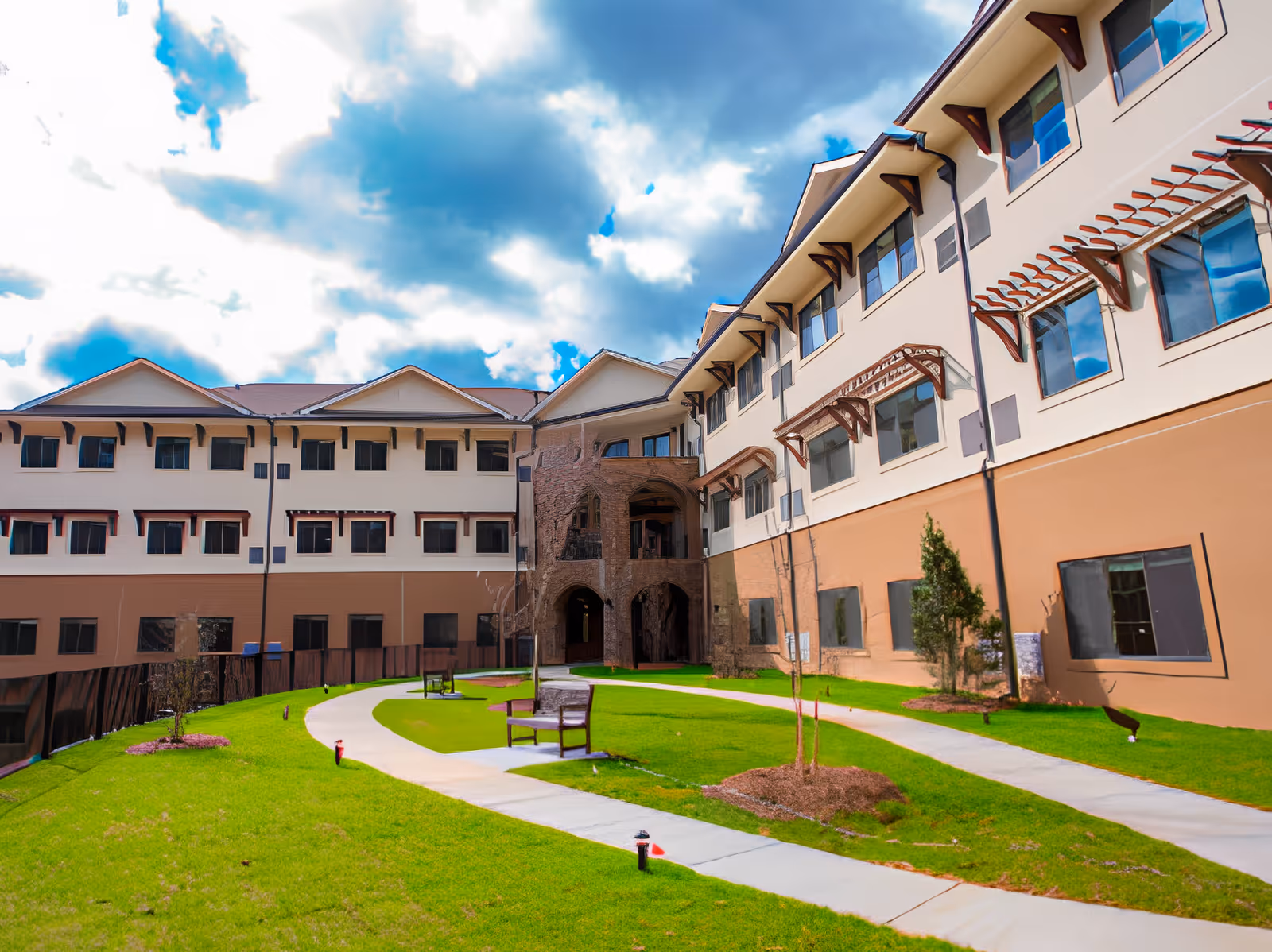 Outdoor courtyard area of Tapestry House Assisted Living at Alpharetta featuring a green lawn with a curved concrete walkway, benches, small trees, and a three-story building with multiple windows surrounding the courtyard under a partly cloudy sky.