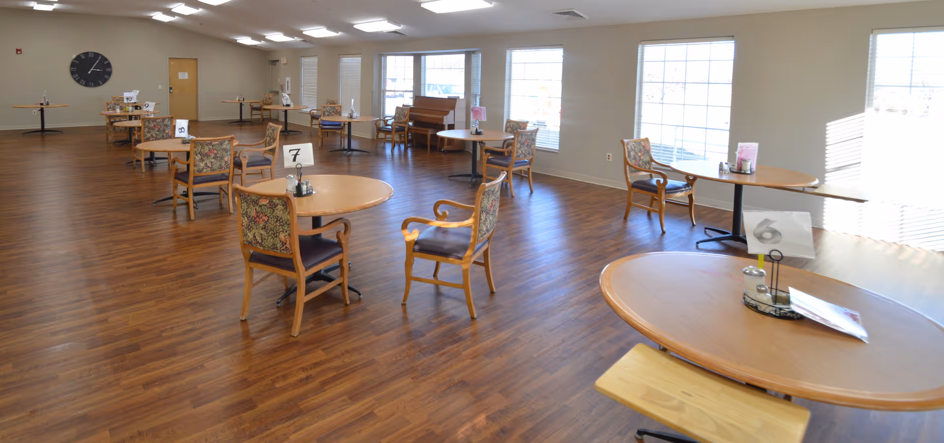 A spacious dining room with multiple round wooden tables and floral upholstered chairs arranged throughout. Each table has a small number card and a condiment set. Large windows on one side let in natural light, and there is a piano against the far wall. The floor is wood laminate, and the walls are painted a light beige color.