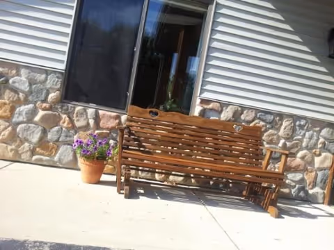Wooden bench on a concrete patio in front of a building with stone and siding exterior walls. A potted plant with purple flowers is placed next to the bench.