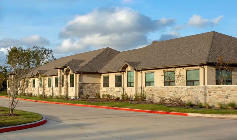 Single-story stone-and-stucco building with multiple windows, a driveway and landscaped curb under a partly cloudy sky.