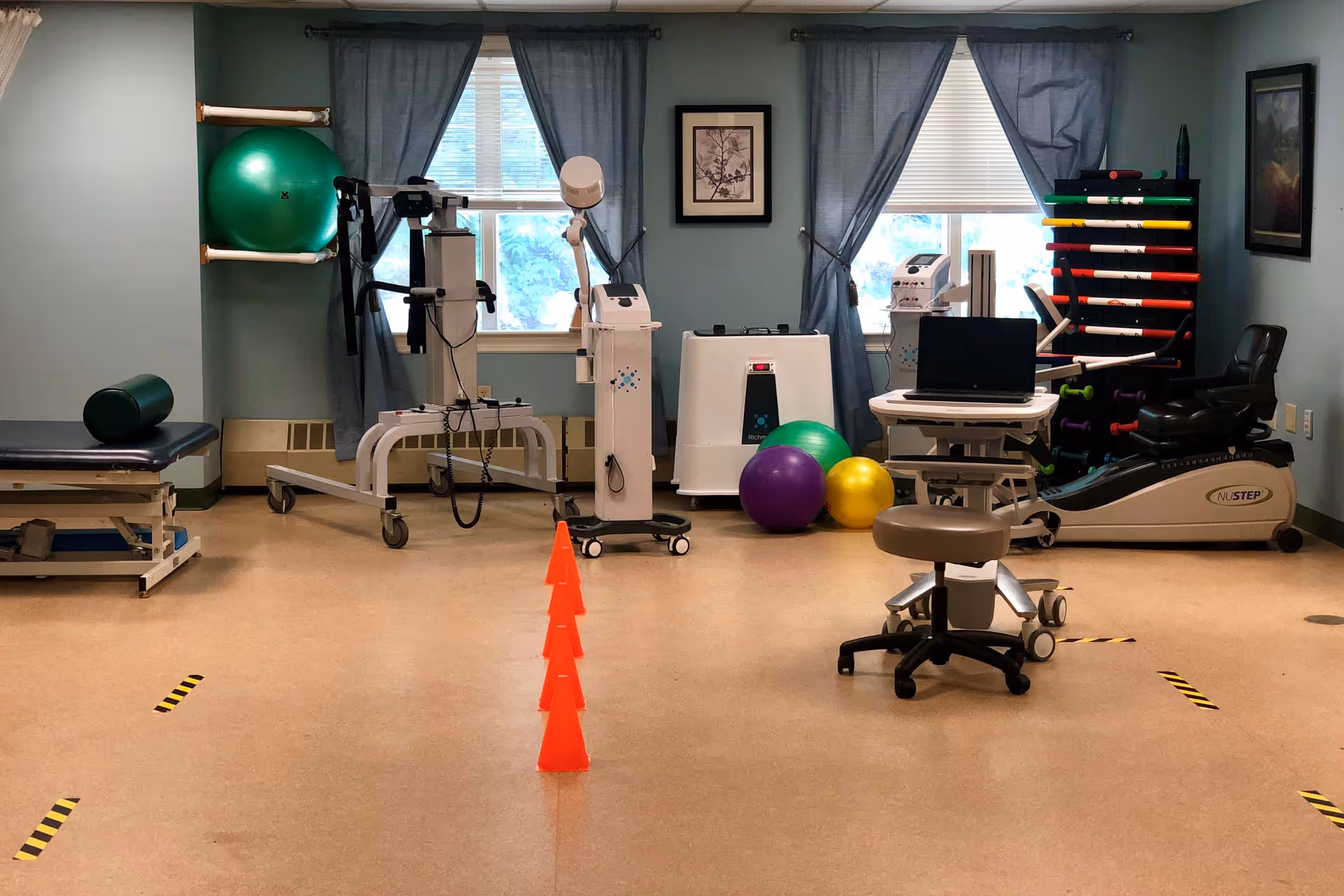 Physical therapy room with exercise machines, therapy balls, a treatment table, and a line of orange cones.