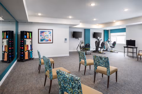 A fitness room with exercise equipment including stationary bikes and a treadmill, several patterned chairs arranged in rows, a rack of colorful dumbbells and exercise bands, two wall-mounted televisions, and a large mirror on one wall. The room is well-lit with recessed ceiling lights and has a window letting in natural light.