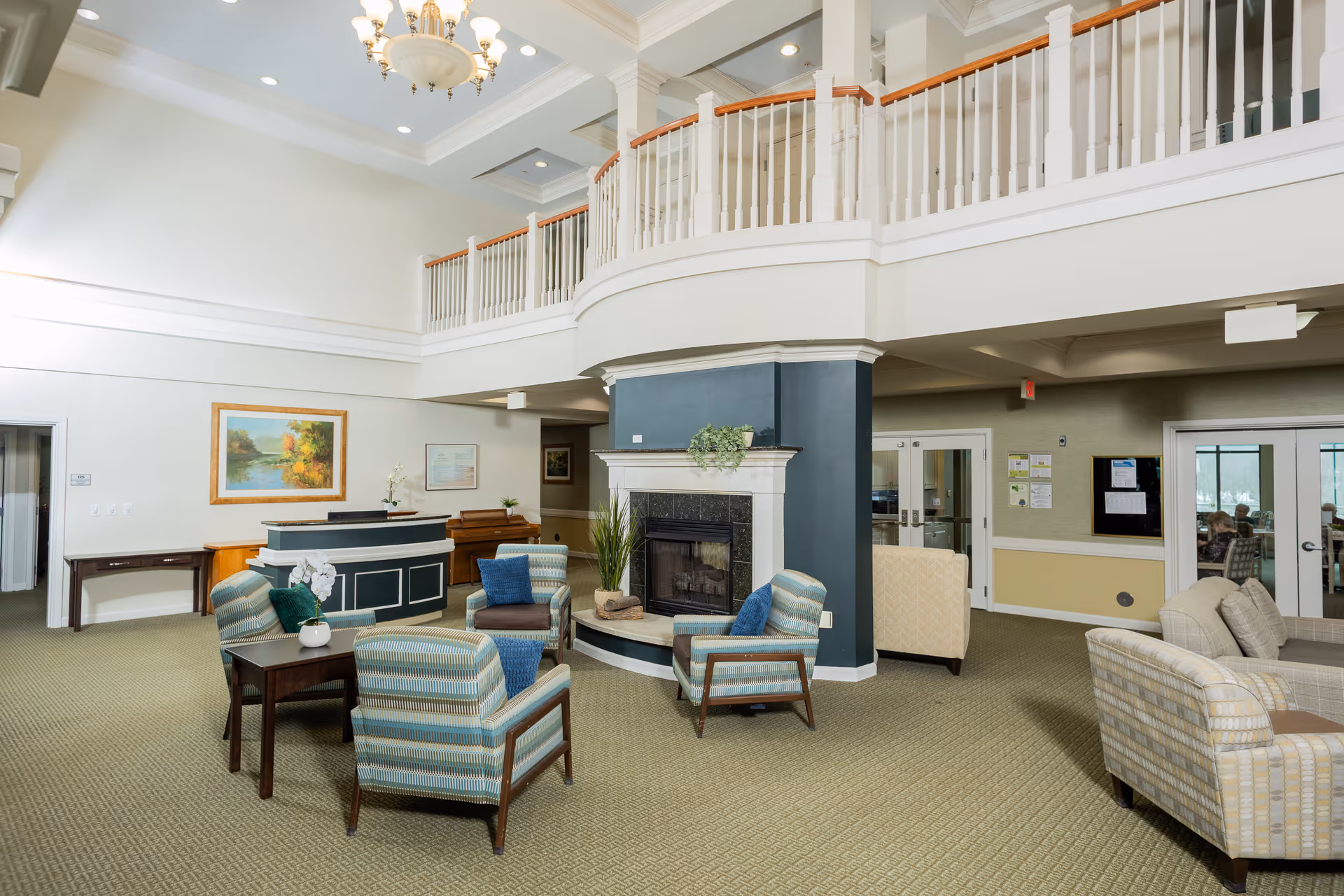 A spacious senior living community common area with a high ceiling and chandelier. The room features a central fireplace with plants on the mantel, surrounded by four striped armchairs with blue cushions and a small wooden table with a white orchid. There is a reception desk in the background, a piano, framed artwork on the walls, and a balcony railing above. Additional seating with beige patterned sofas is visible on the right side near glass doors.