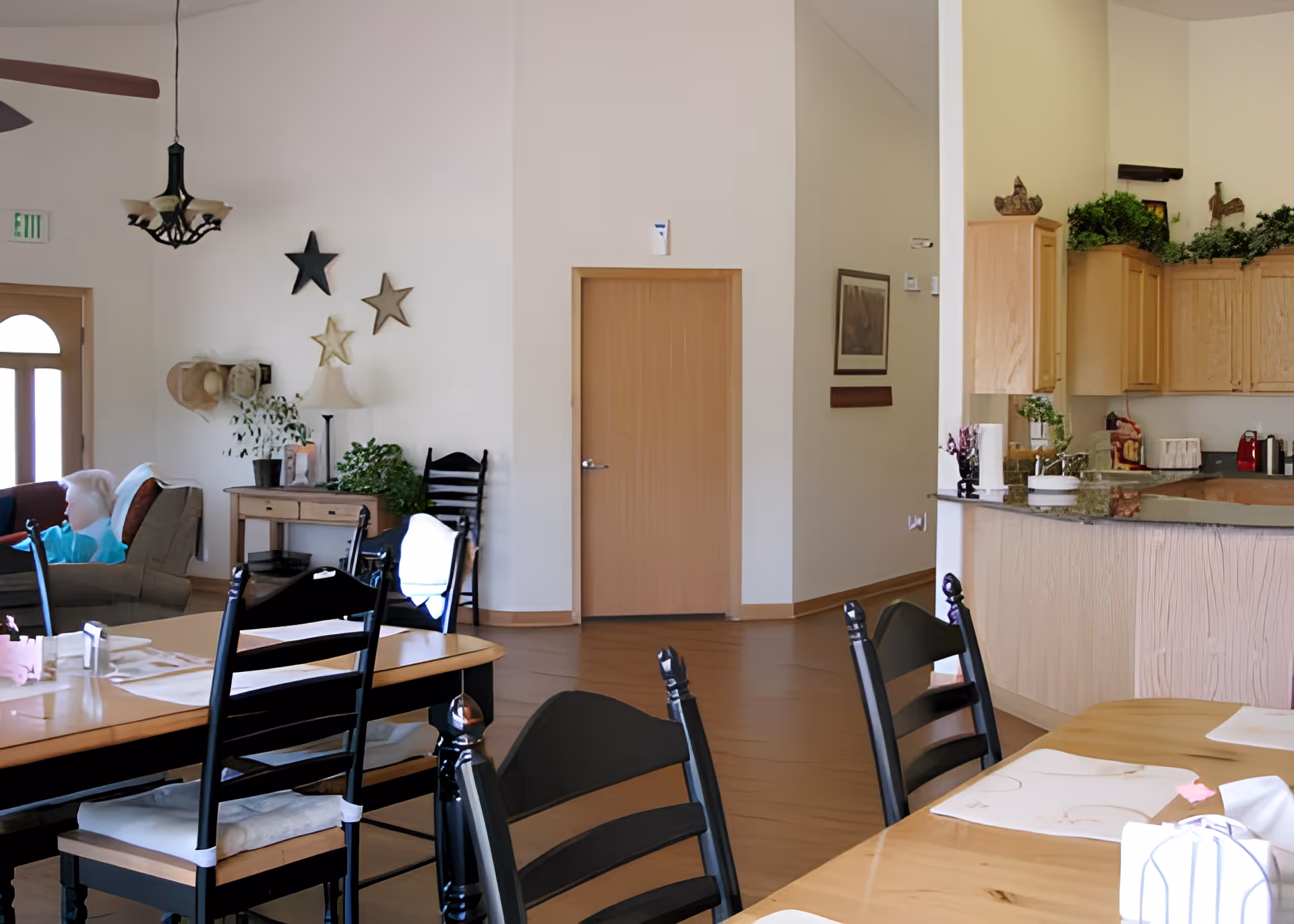 Communal dining area and open kitchen in a senior living facility with tables, chairs, and a seating area.