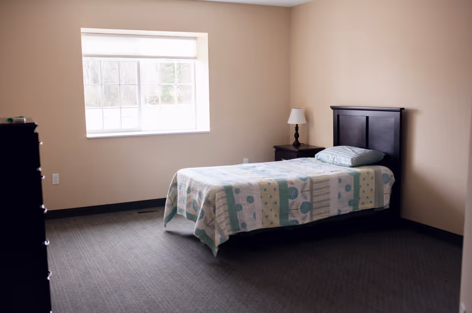 A simple bedroom with a single bed covered with a patterned quilt, a pillow, a wooden headboard, a nightstand with a lamp, a window letting in natural light, and a dresser partially visible on the left side.
