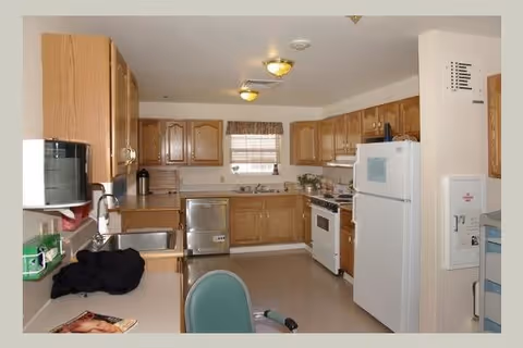 Interior view of a kitchen with wooden cabinets, a white refrigerator, a white stove, a dishwasher, a microwave, and a sink. There is a window with blinds above the sink and a green chair partially visible in the foreground.