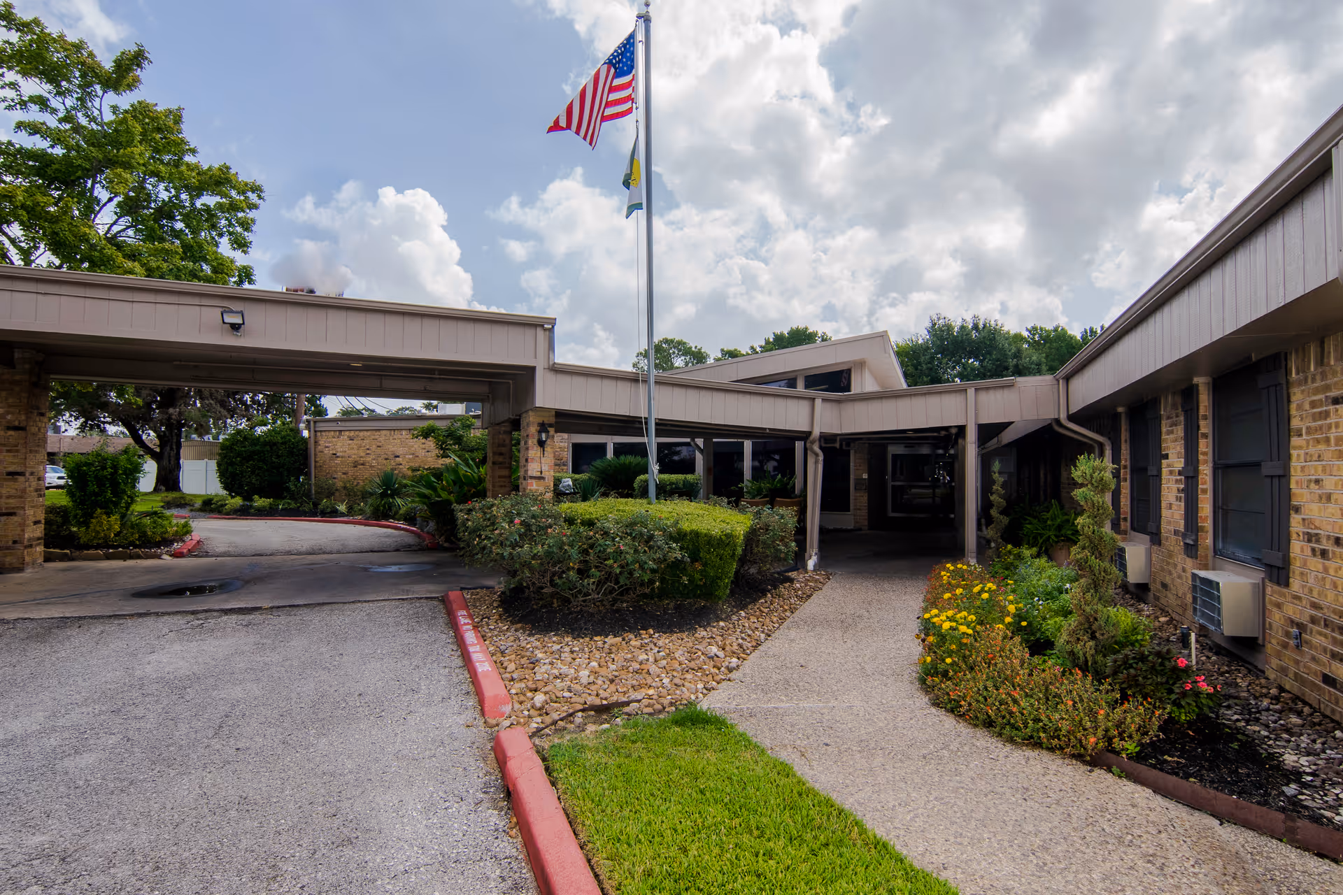 Entrance driveway and covered drop-off with a flagpole and landscaped walkway in front of a one-story brick senior care facility.