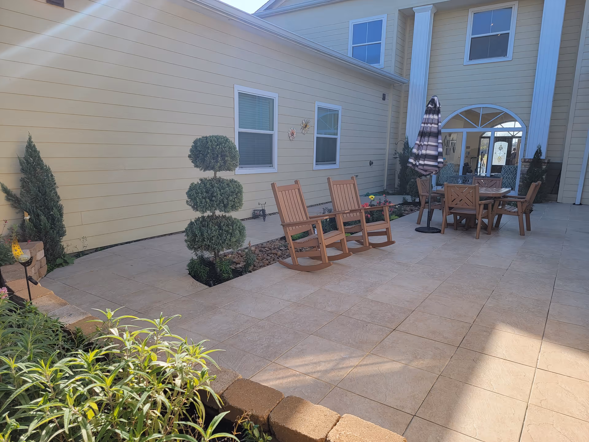 Outdoor patio area with tiled flooring, two wooden rocking chairs, a round wooden table with four chairs, and a closed striped umbrella. The patio is adjacent to a yellow building with white trim and two windows. There are decorative plants and small trees along the edge of the patio.