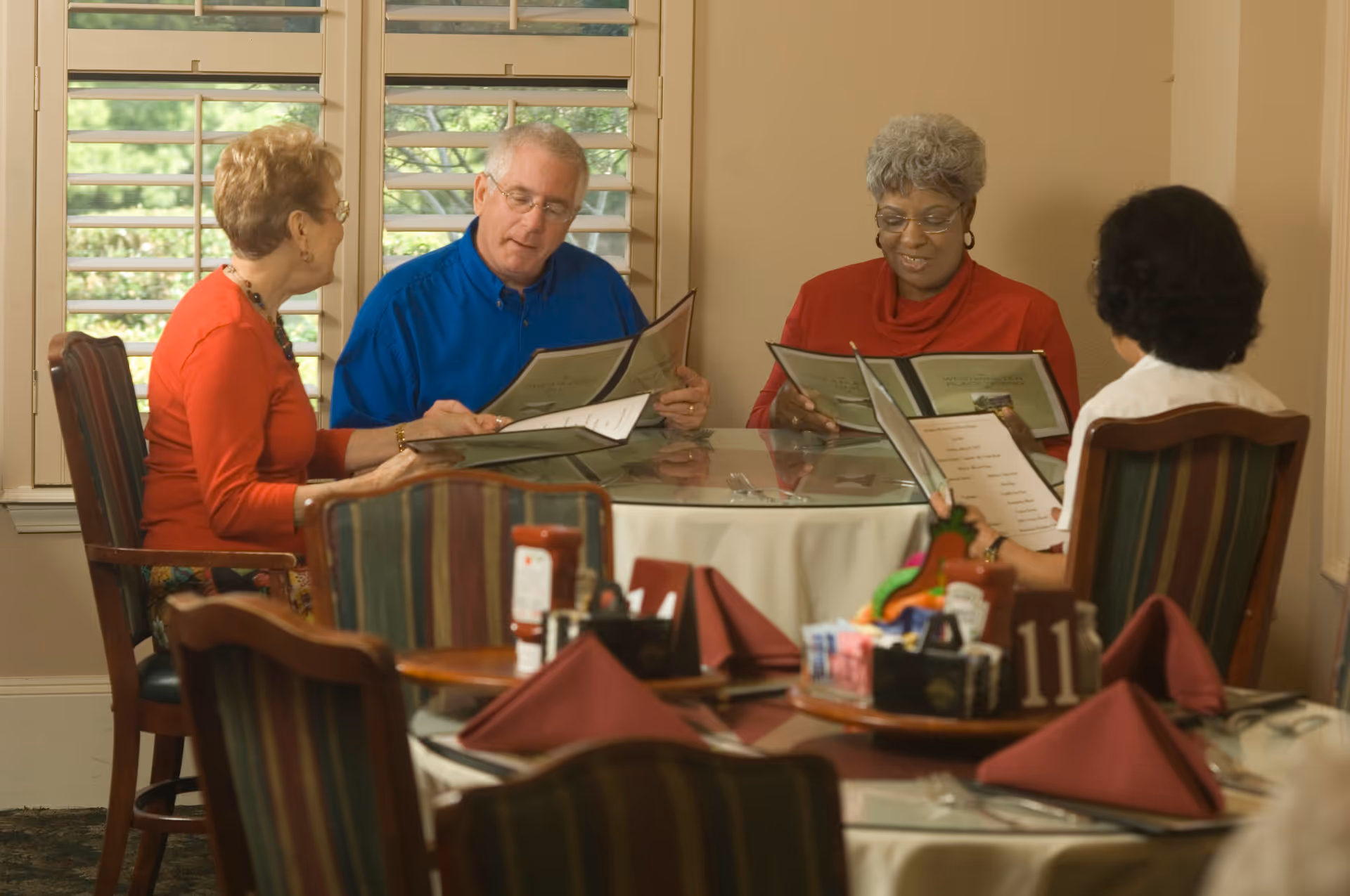Four older adults seated around a round dining table reading menus in a dining room.