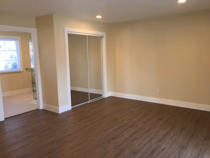 Empty room with wood flooring, beige walls, recessed ceiling lights, a mirrored sliding closet door, and an open doorway leading to a bathroom with a window and tiled floor.