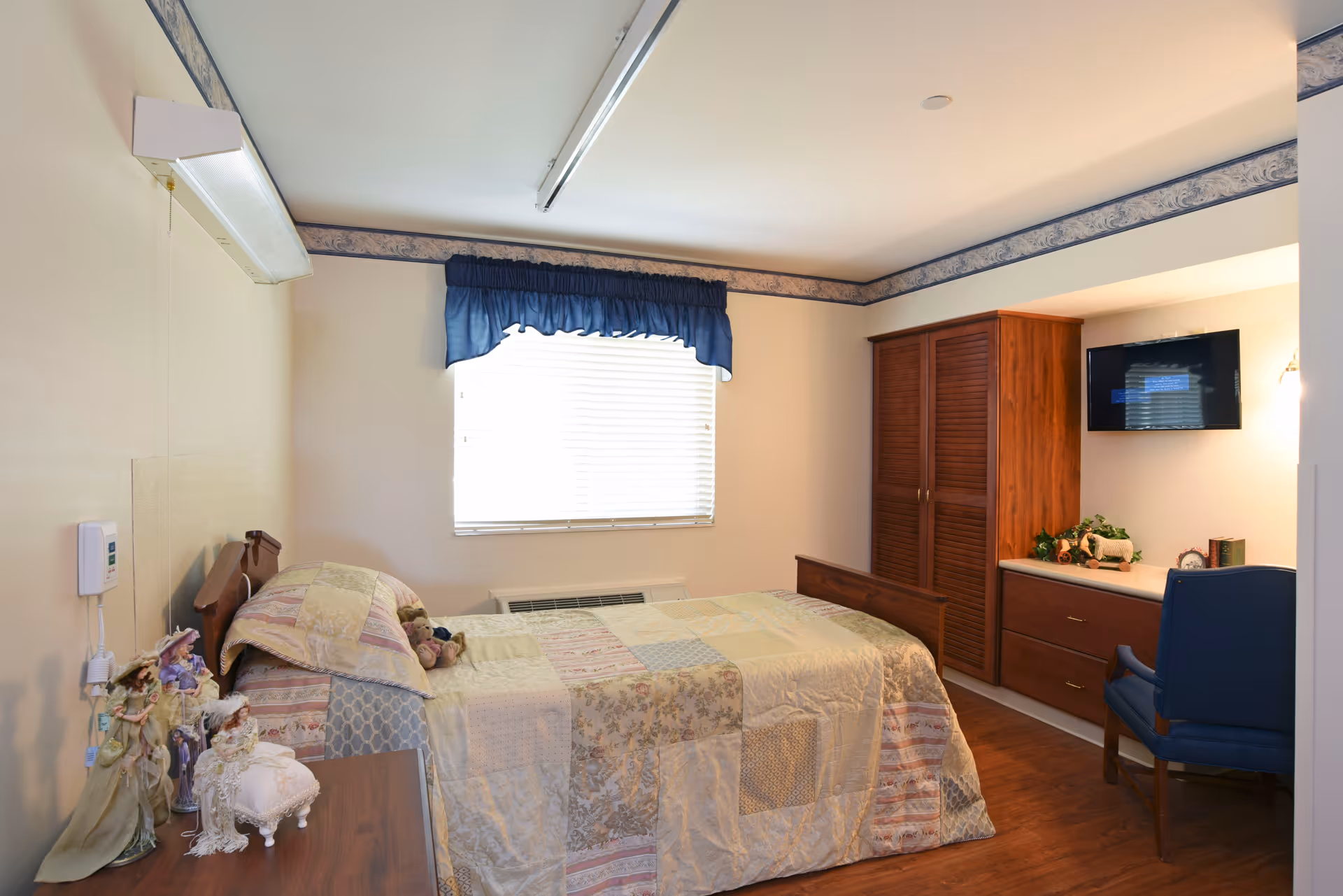 A cozy bedroom in a nursing facility featuring a single bed with a patchwork quilt, a wooden headboard, and stuffed animals on the bed. There is a window with white blinds and a blue valance, a wooden dresser with decorative items, a blue chair, a mounted flat-screen TV, and a wooden wardrobe. The room has light-colored walls and wood flooring.