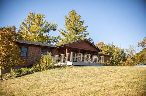 A single-story brick building with a covered porch and railing, surrounded by trees and a grassy hill under a clear blue sky.