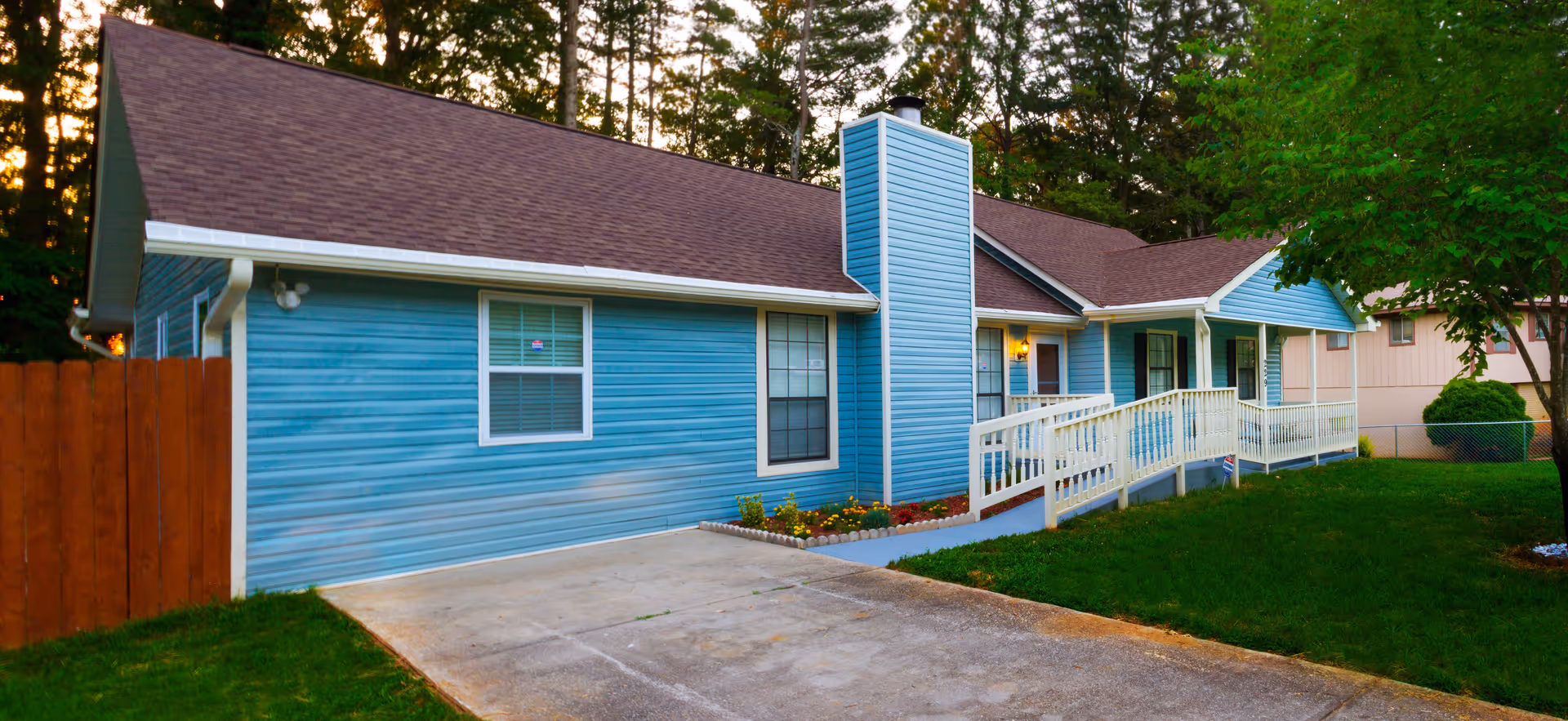 Exterior view of a single-story residential building with blue siding, a brown shingled roof, a white railing ramp leading to the front door, and a concrete driveway. The building is surrounded by green grass, trees, and a wooden fence on the left side.
