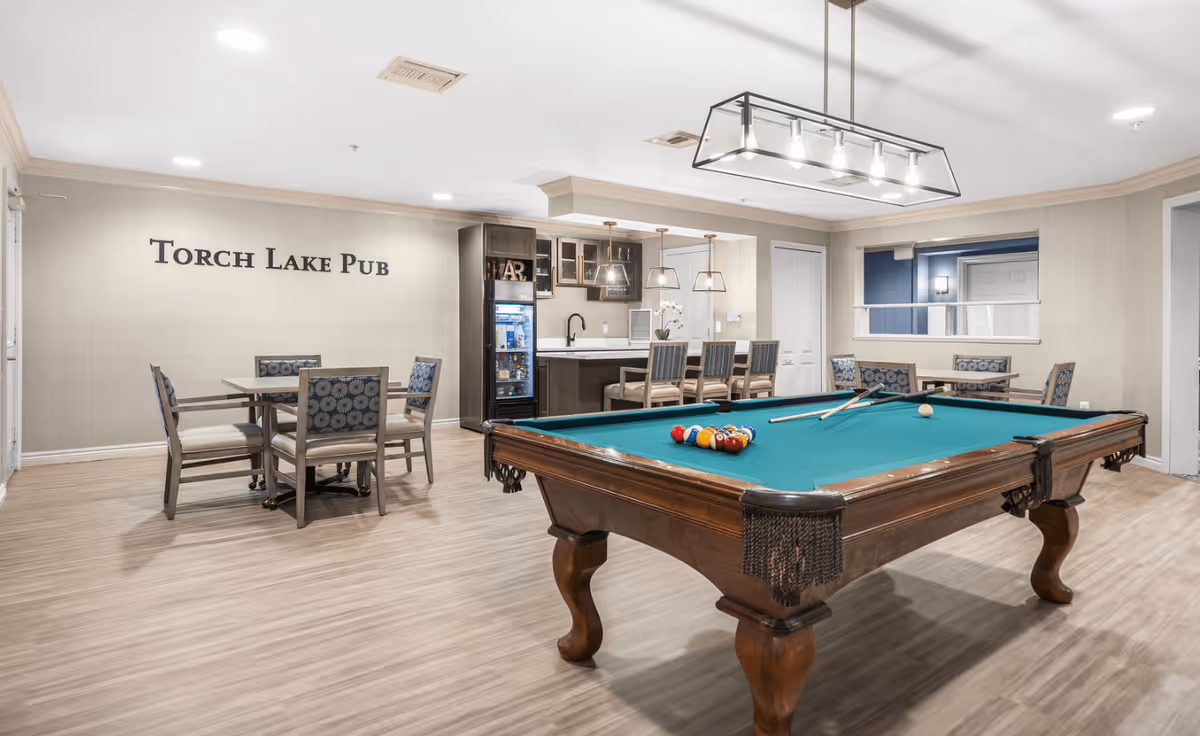 A recreational room named Torch Lake Pub featuring a pool table with balls and cues, a seating area with tables and chairs, and a small bar with high chairs and pendant lights. The room has light-colored walls and wood flooring.