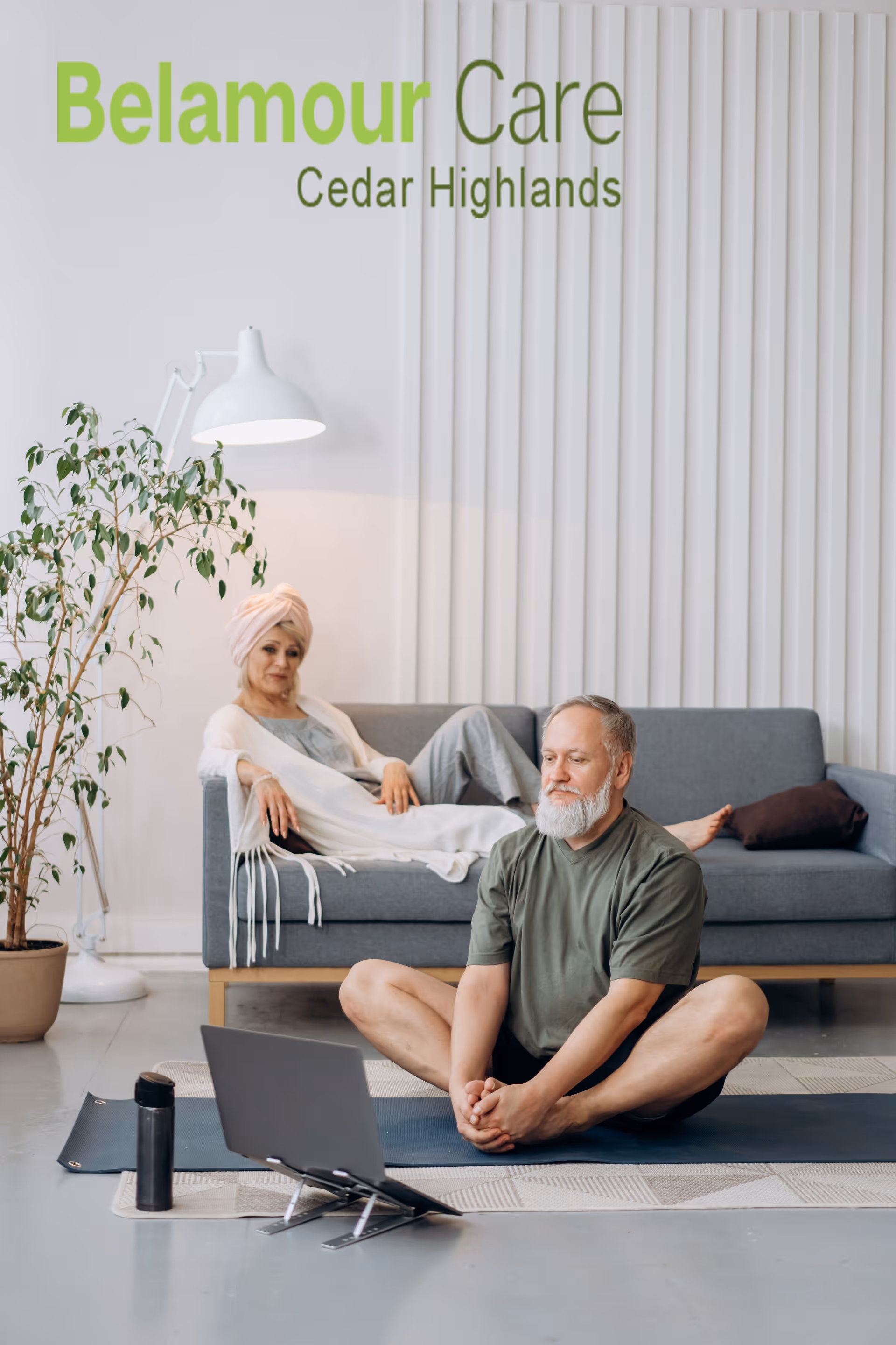 An elderly man is sitting on a yoga mat on the floor in a butterfly stretch position, looking at a laptop in front of him. Behind him, an elderly woman with a head wrap is relaxing on a gray sofa. The room has a modern, minimalist design with a potted plant and a white floor lamp. The text 'Belamour Care Cedar Highlands' is displayed on the wall above them.