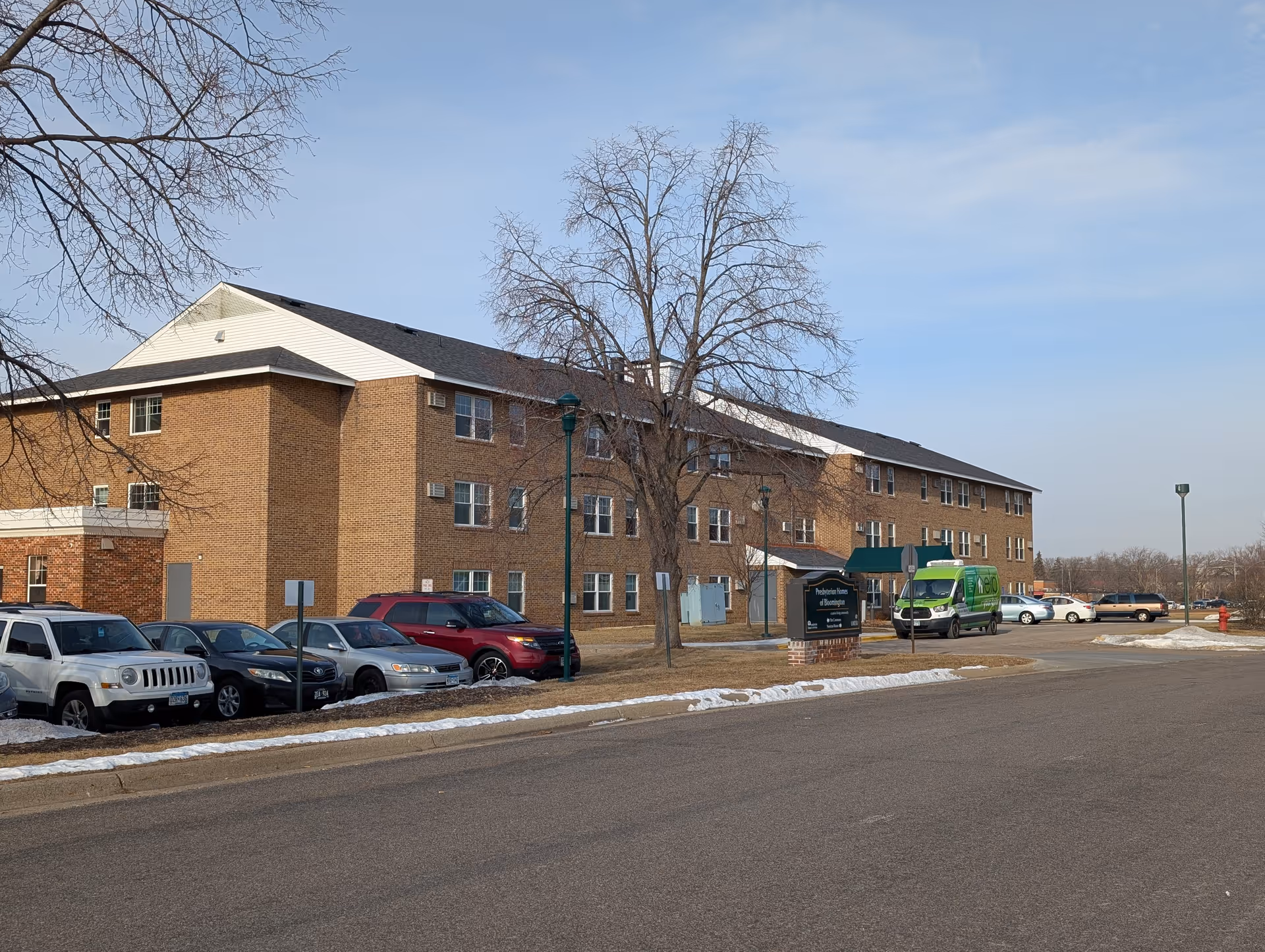 Three-story brick senior living building with parked cars and a sign in front on a clear day.