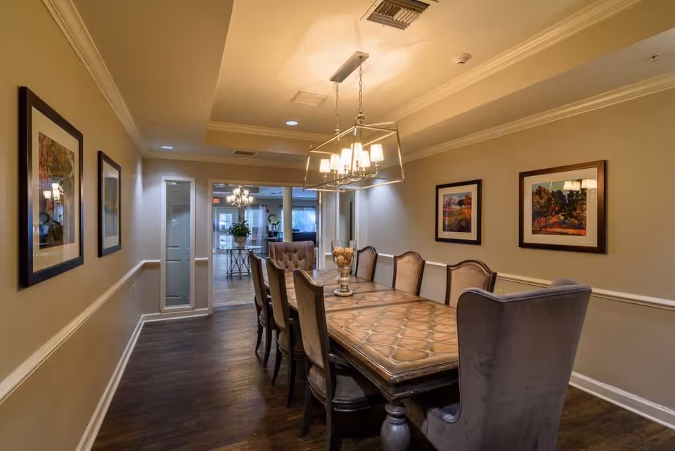 A well-lit dining room with a long wooden table surrounded by upholstered chairs. The room has beige walls adorned with framed artwork and a modern chandelier hanging above the table. The floor is dark wood, and there is an open doorway leading to another room with more seating and natural light.