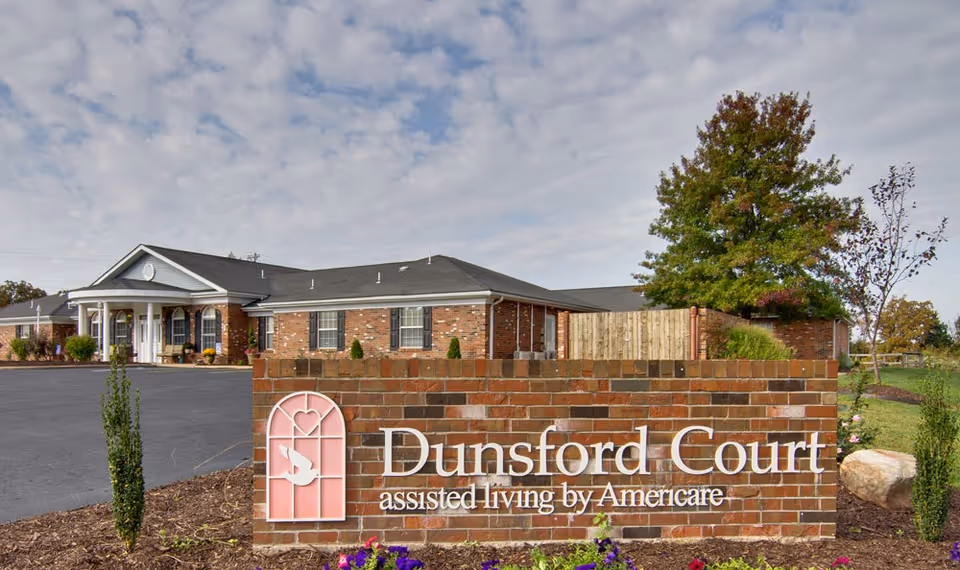 Exterior view of a brick building with a sign in front that reads 'Dunsford Court assisted living by Americare'. The building has a dark roof, white columns at the entrance, and is surrounded by a paved driveway and landscaping with trees and shrubs under a partly cloudy sky.