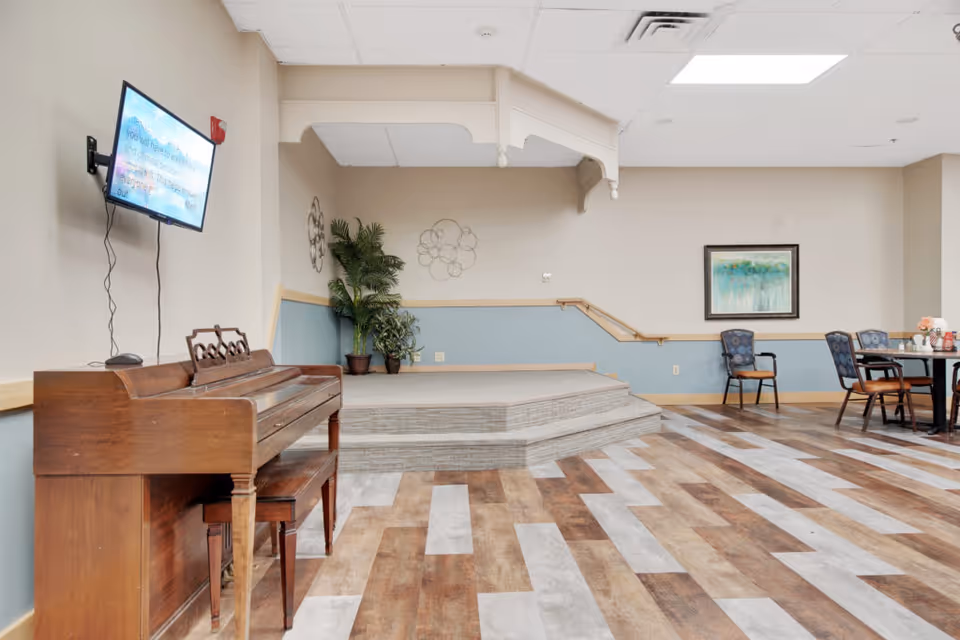 Community activity room with a wooden piano, a small raised stage, wall-mounted TV, chairs and tables, and potted plants.