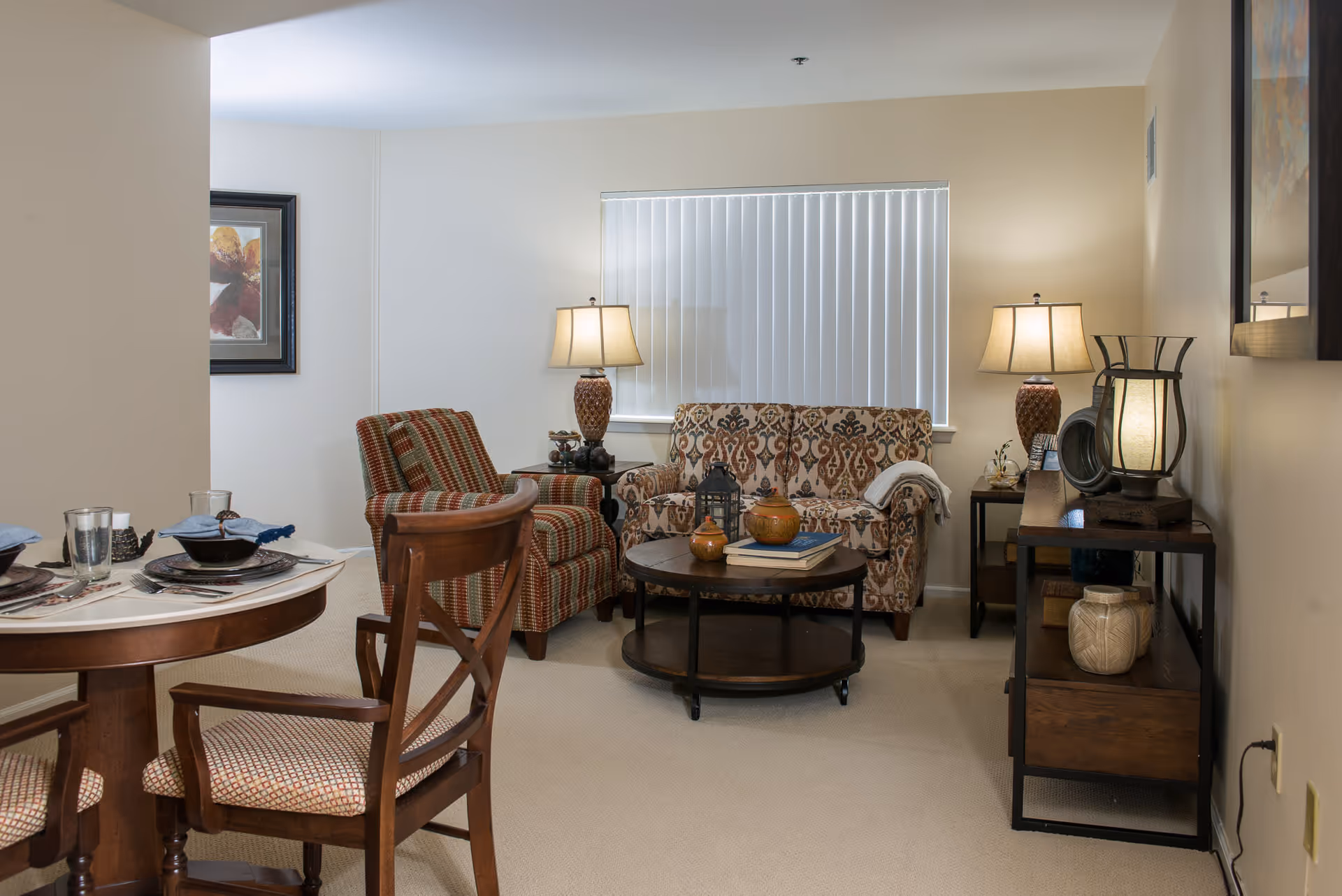 A cozy living room area with a patterned sofa, a striped armchair, two table lamps, a round wooden coffee table with decorative items, and a wooden console table with lamps and vases. Adjacent to the living area is a dining table set with plates, bowls, glasses, and napkins. The room has beige walls and carpet, and a window with vertical blinds.