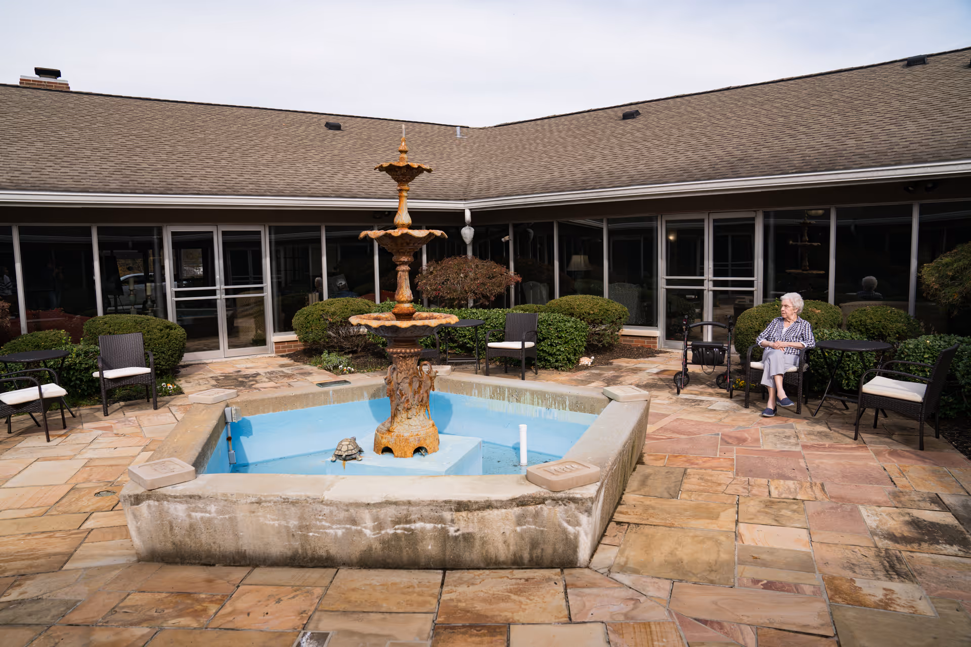 Outdoor courtyard area at Village At Wesley Manor featuring a stone-paved patio with a central multi-tiered fountain. There are several black metal tables and chairs with cushions arranged around the courtyard. An elderly woman is seated at one of the tables near some green bushes and a walker. The building with large windows and a sloped roof surrounds the courtyard.