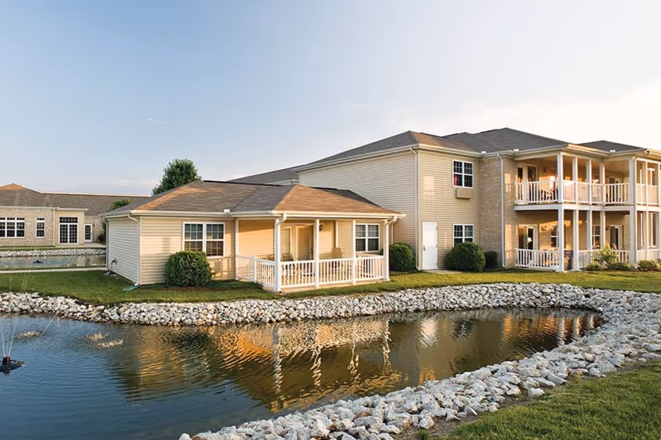 Two-story residence and a one-story unit with covered porches reflected in a pond bordered by rocks.