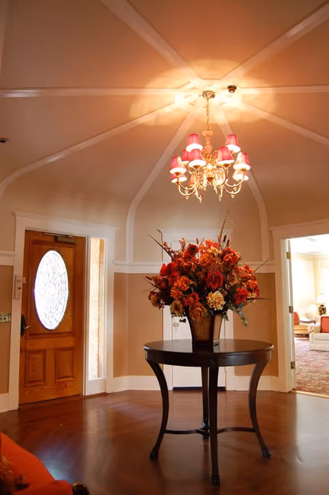A warmly lit foyer with a round table holding a large floral arrangement, a chandelier overhead, and a wooden front door to the left.