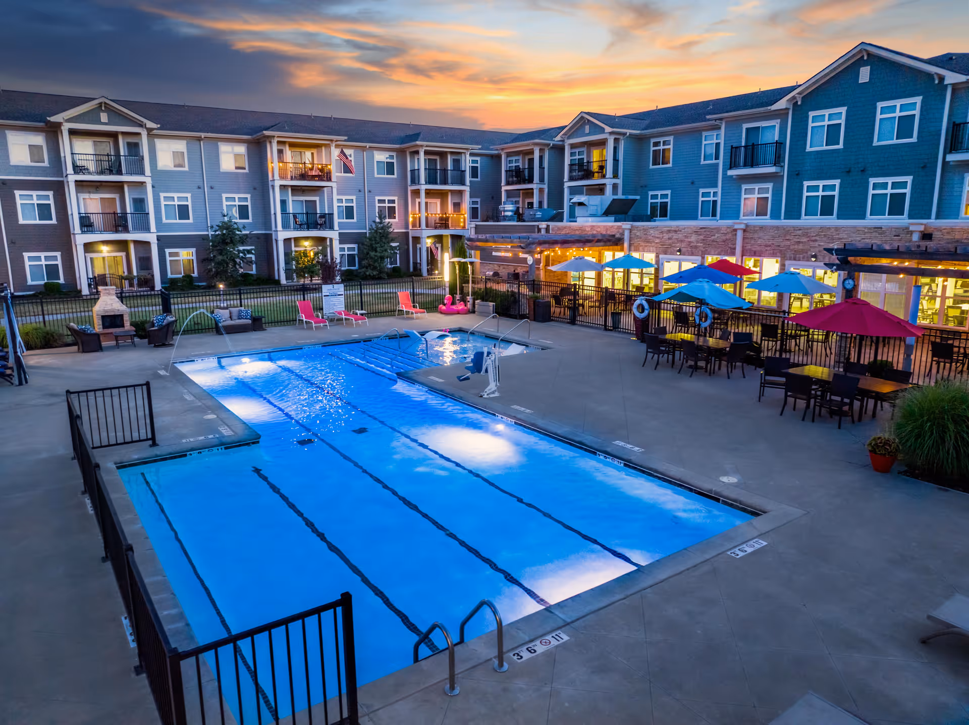 Outdoor swimming pool area at a senior living facility during sunset, surrounded by a three-story residential building with balconies. The pool is illuminated with underwater lights and has a pool lift for accessibility. There are lounge chairs, colorful umbrellas, and tables on the pool deck, along with some greenery and a fireplace in the background.