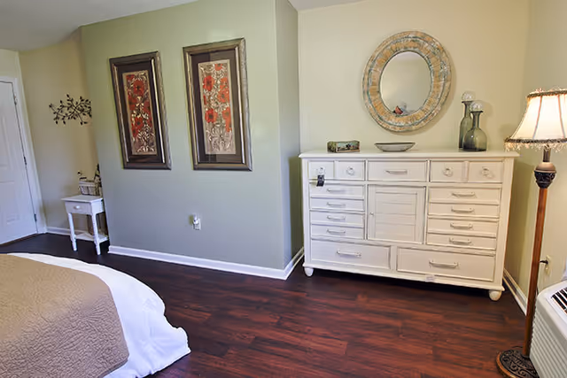 A bedroom with dark wood flooring, a white dresser with multiple drawers, a round decorative mirror above the dresser, two framed floral artworks on the wall, a small white side table with a plant, and a floor lamp with a beige lampshade. Part of a bed with white and beige bedding is visible in the foreground.
