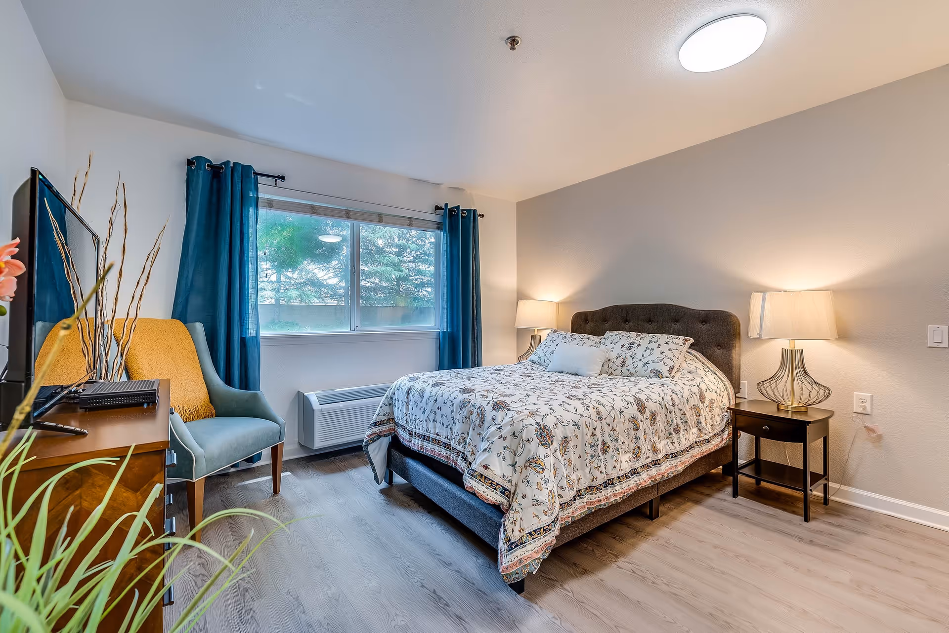 A bright and cozy bedroom with a large window covered by blue curtains. The room features a bed with a floral patterned comforter and pillows, two bedside tables each with a lamp, a wooden dresser with a TV on top, and a comfortable armchair with a yellow throw blanket. The floor is light wood, and the walls are painted in neutral tones.