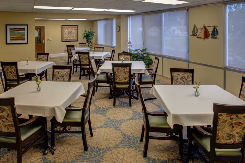 A dining room with several square tables covered with white tablecloths, each table surrounded by wooden chairs with floral-patterned cushions. The room has large windows with blinds, a carpet with a floral pattern, and wall decorations including a framed picture and a wall art of sailboats.