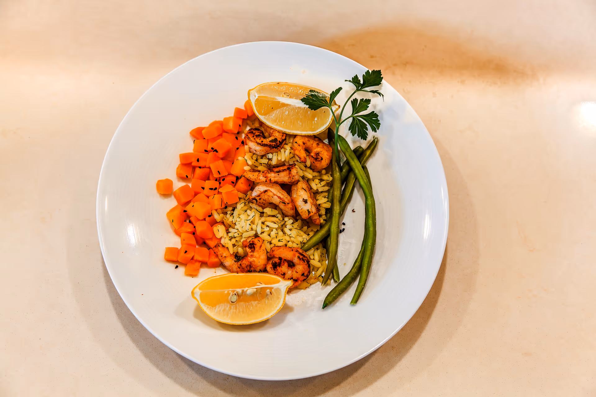 Top-down view of a white plate with rice, grilled shrimp, green beans, diced carrots, and lemon wedges on a light countertop.