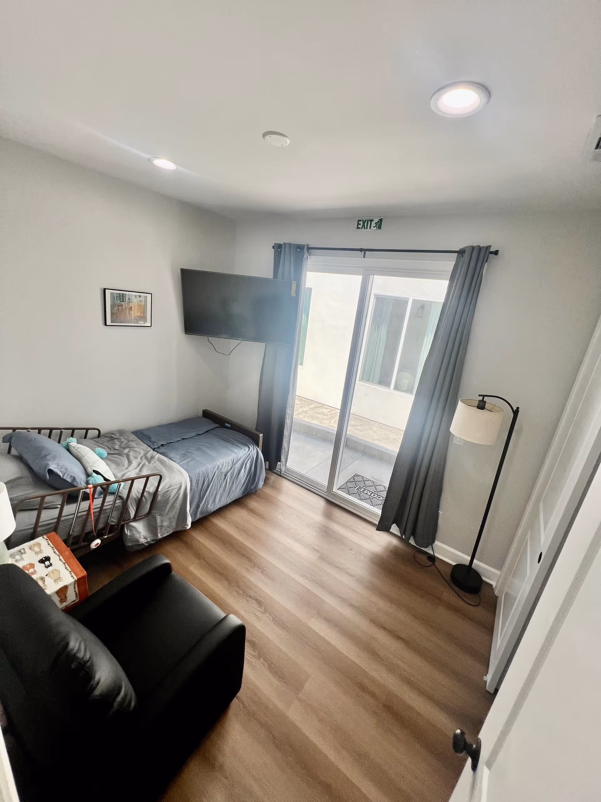 A small bedroom with a single bed covered in gray bedding, a black recliner chair, a floor lamp, a wall-mounted flat-screen TV, and sliding glass doors with gray curtains leading to an outdoor area. The room has wood flooring and light gray walls with a small framed picture and an exit sign above the door.