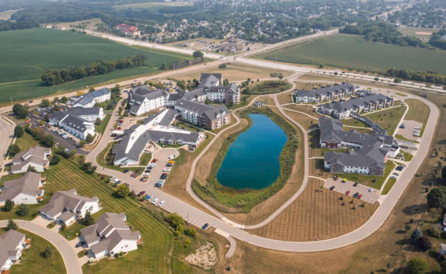 Aerial view of The Homestead at Rochester senior living campus with multiple buildings surrounding a central pond and roadways.