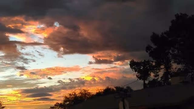 A dramatic sunset sky with dark clouds and patches of orange, yellow, and blue light above silhouetted trees and rooftops.