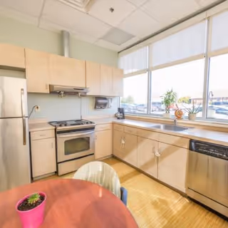 Bright kitchen area with light wood cabinets, stainless steel refrigerator, oven, and dishwasher. A round wooden table with a small potted plant in a pink pot is in the foreground. Large windows with white blinds let in natural light and show a parking lot outside.