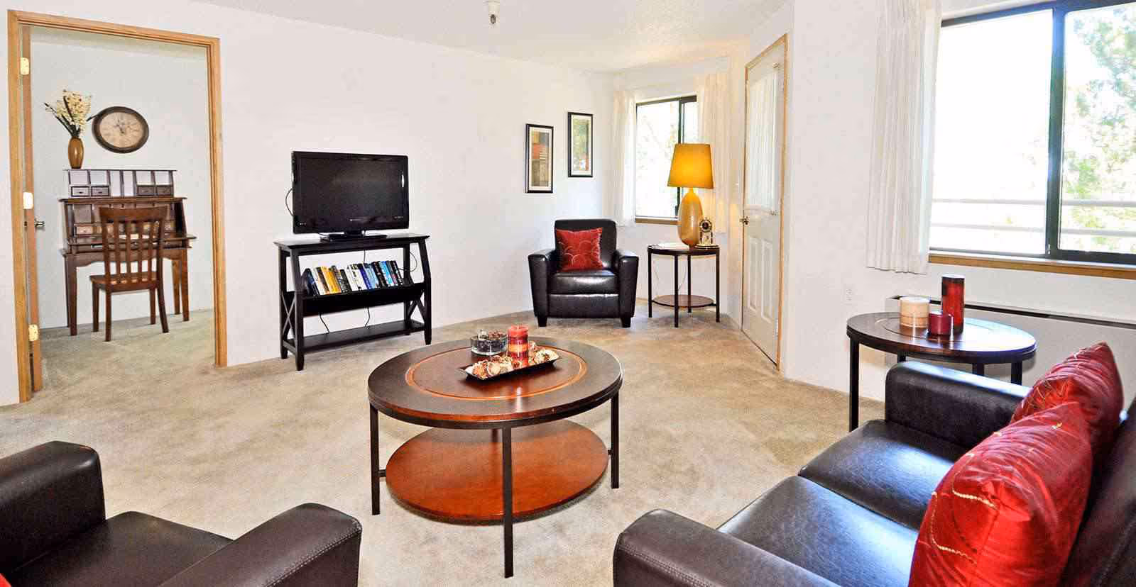 A bright living room with beige carpet, a black leather sofa with red pillows, two black leather armchairs, a round wooden coffee table with candles and decorative items, a TV on a black stand with books, a side table with a lamp, and a window with white curtains. An adjoining room with a wooden desk, chair, and wall clock is visible through an open doorway.