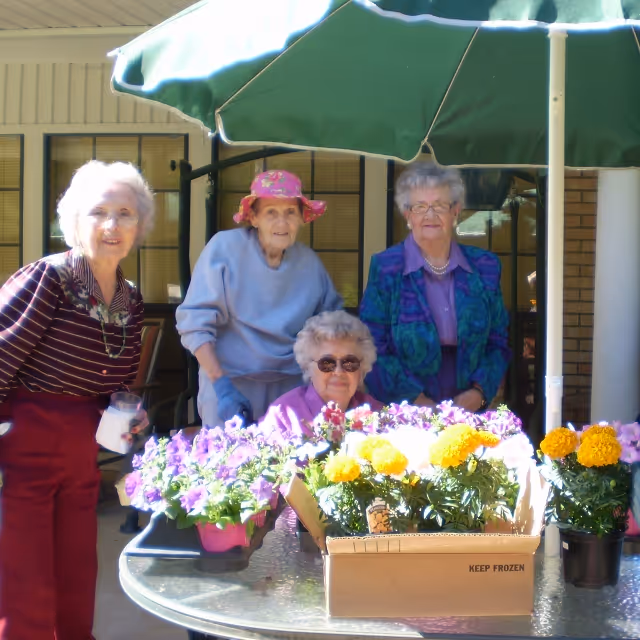 Four elderly women gathered around a round outdoor table with colorful potted flowers and a cardboard box. They are under a large green patio umbrella, with a building and windows in the background. One woman is wearing a pink floral hat, and another is holding a glass of milk.