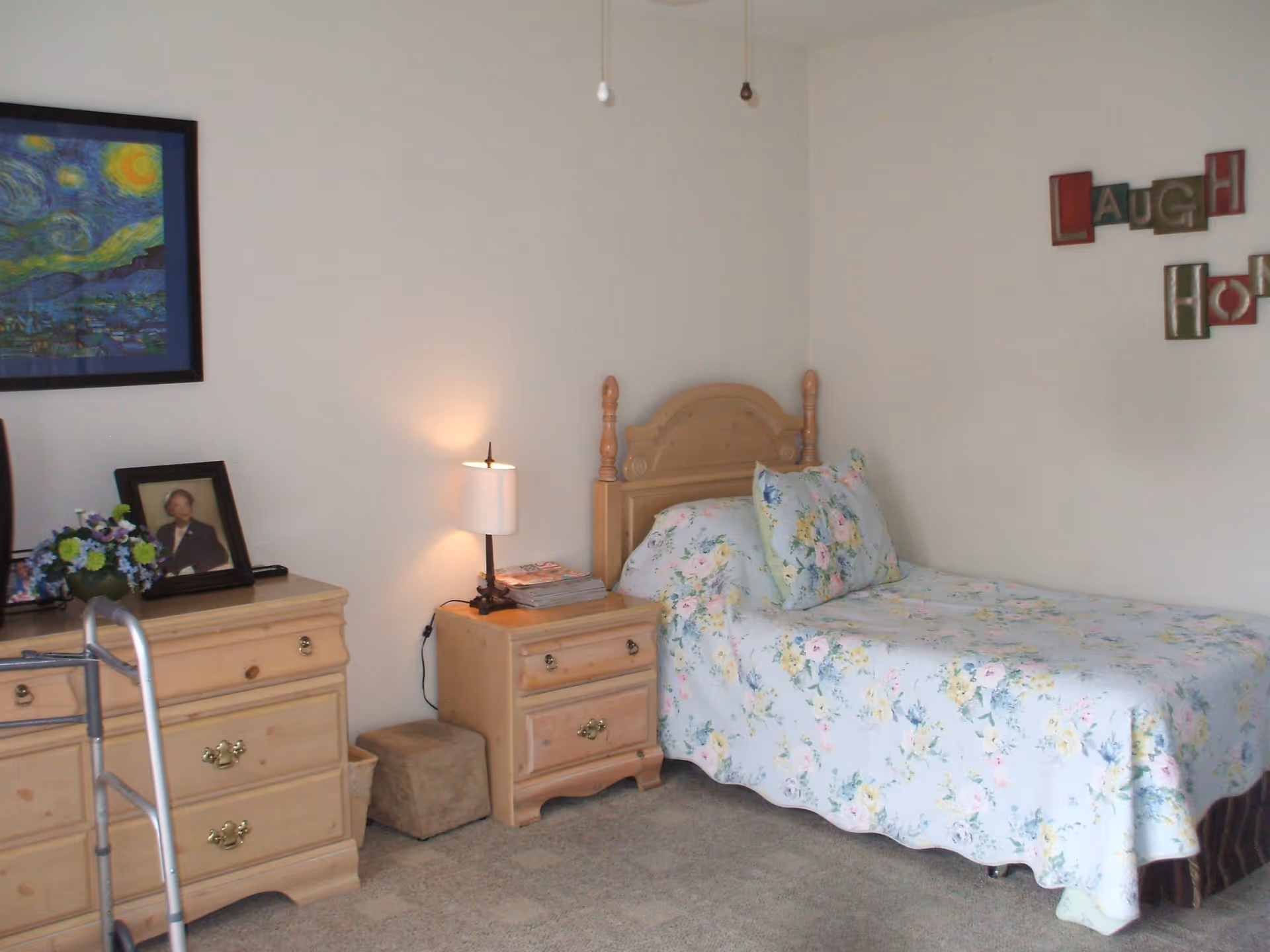 A bedroom with a single wooden bed covered with a floral bedspread and matching pillow. Next to the bed is a wooden nightstand with a small lamp and some magazines. There is a wooden dresser with a framed photograph and a vase of flowers on top. A walker is partially visible in front of the dresser. The walls are plain with a framed painting and decorative wall art spelling 'LAUGH' and 'HOME'.
