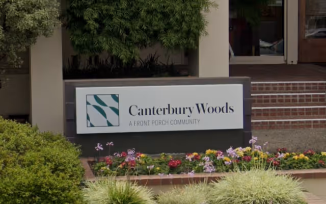 Outdoor view of a sign for Canterbury Woods, a front porch community, surrounded by greenery and flowers with a building entrance in the background.