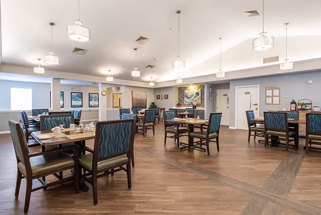 A spacious dining room with multiple wooden tables and chairs featuring blue patterned upholstery. The room has wood flooring, modern pendant lights hanging from the ceiling, and artwork on the walls. There is a fireplace with a painting above it in the background.