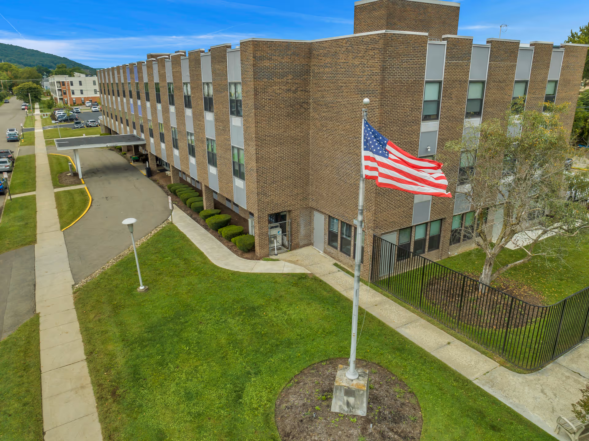 Exterior view of a three-story brick building with multiple windows, a covered entrance driveway, a sidewalk, and a flagpole with an American flag waving in front. The building is surrounded by green grass, bushes, and a tree, with a street and parked cars visible on the left side.