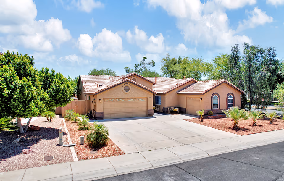 Exterior view of a single-story residential building with a tiled roof, two garages, and a driveway. The front yard is landscaped with small palm trees, bushes, and red mulch. The sky is partly cloudy with blue patches.
